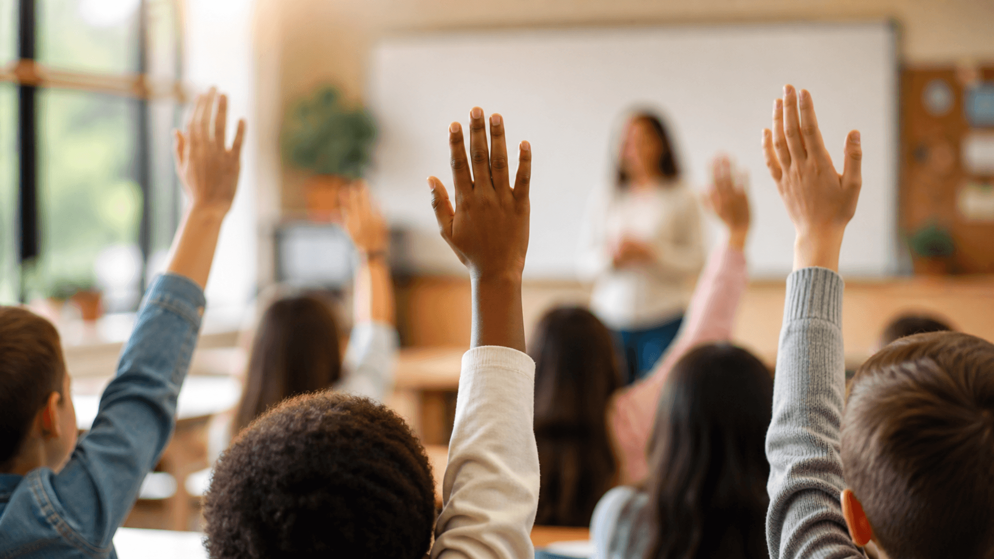 Students raising their hands in a classroom while a teacher stands at the front of the room